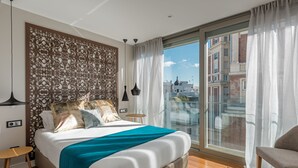 A modern hotel room with a large bed, a decorative headboard, a view of the city through a glass door, and a balcony with a chair. - Gran View Apartments (Madrid)