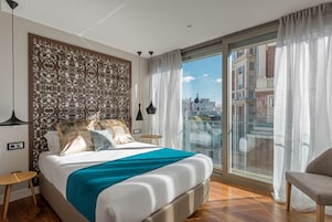 A modern hotel room with a large bed, a decorative headboard, a view of the city through a glass door, and a balcony with a chair.