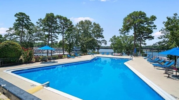 Une piscine rectangulaire à l’eau bleue, entourée de chaises longues et de parasols, avec des arbres et une étendue d’eau visible au loin.