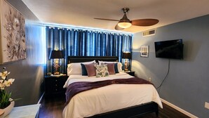 Une chambre à coucher comprenant un lit, des tables de chevet, un ventilateur de plafond, un téléviseur et une fenêtre avec des rideaux.