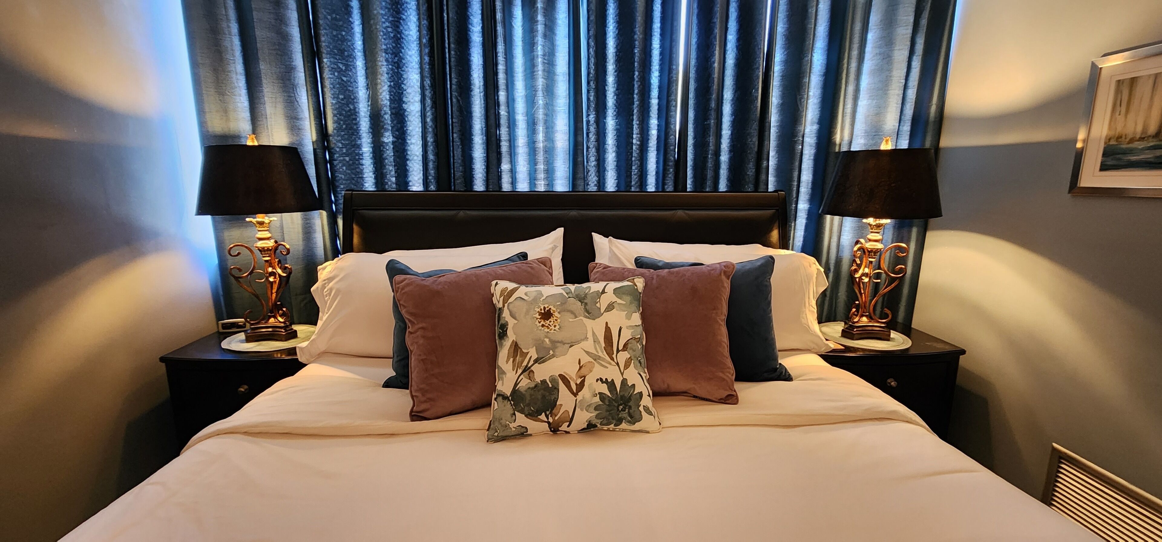 A neatly made bed with a floral pillow, two side tables with lamps, and a view of the night sky through blue curtains.