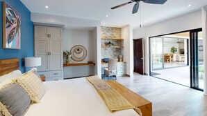 Une chambre moderne avec un lit, une table de chevet et un ventilateur de plafond. Une pièce adjacente contient un bureau et une chaise, et une grande fenêtre offre une vue sur l’extérieur.