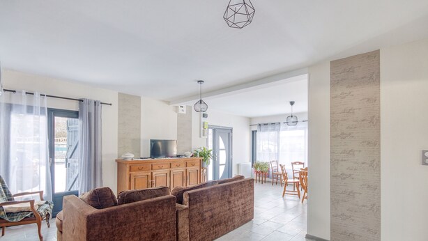 A modern living room with a brown sectional sofa, wooden dining table, and chairs. There is a kitchen area with a counter and cabinets, and a large window with curtains.