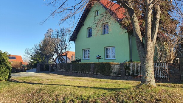 Een groene woning met een rood dak, omringd door een witte hek en een boom.