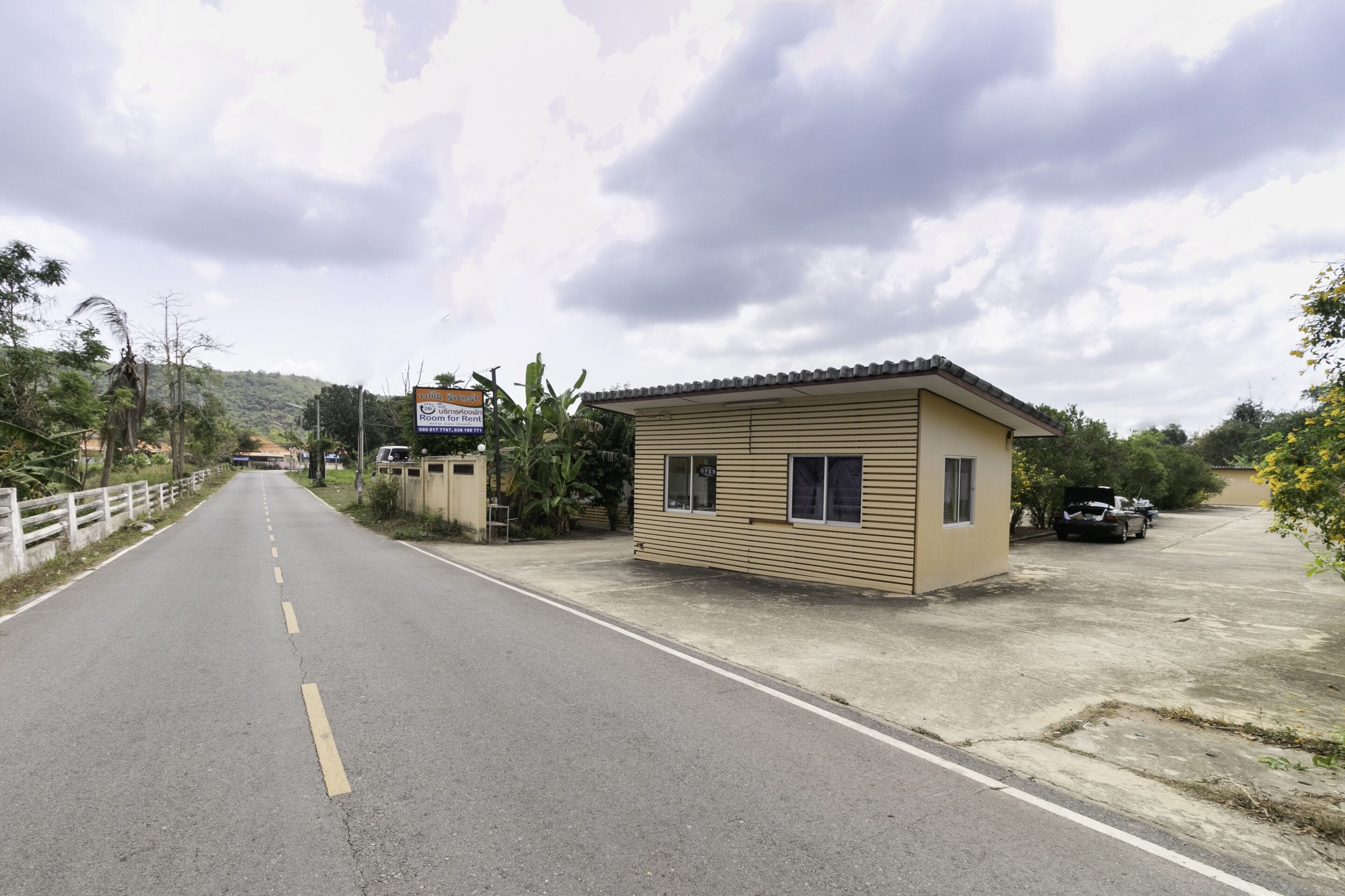 A road leading to a small building with a signboard, surrounded by greenery and a cloudy sky.