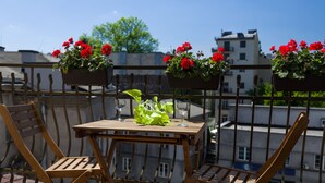 A wooden table with two chairs on a balcony, with red flowers in hanging baskets and a clear view of buildings in the background. - KrakowRentals - Vanilla 5 Apartment (Kraków)