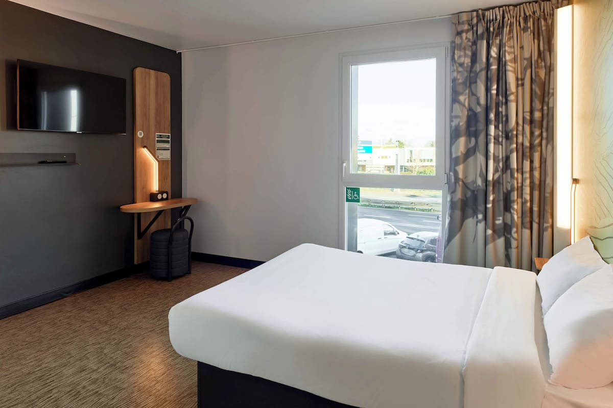 Habitació d'hotel amb un llit gran, una televisió de pantalla plana penjada a la paret, una taula petita amb una làmpada i una finestra amb cortines.