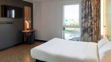 A hotel room with a large bed, a flat-screen TV mounted on the wall, a small table with a lamp, and a window with curtains.
