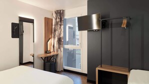 Ein Hotelzimmer mit zwei Betten, einem Schreibtisch, einem Fernseher und einem Fenster mit Vorhängen.
