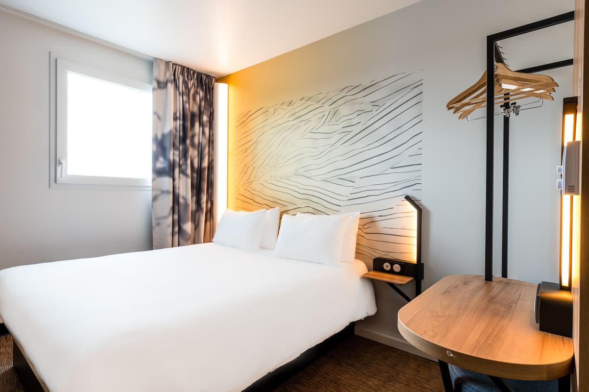 Habitació d'hotel amb un llit gran, una taula de nit amb una làmpada, un penjador de parets i una finestra amb cortines.
