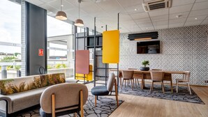 Eine moderne Hotelhalle mit einer Couch, Stühlen, einem Esstisch, Pendelleuchten und einem Fernseher an der Wand.