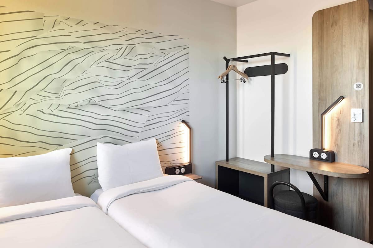Habitació d'hotel amb dos llits, una làmpada penjada a la paret, un escriptori de fusta amb una cadira i una perxa per a abrics.