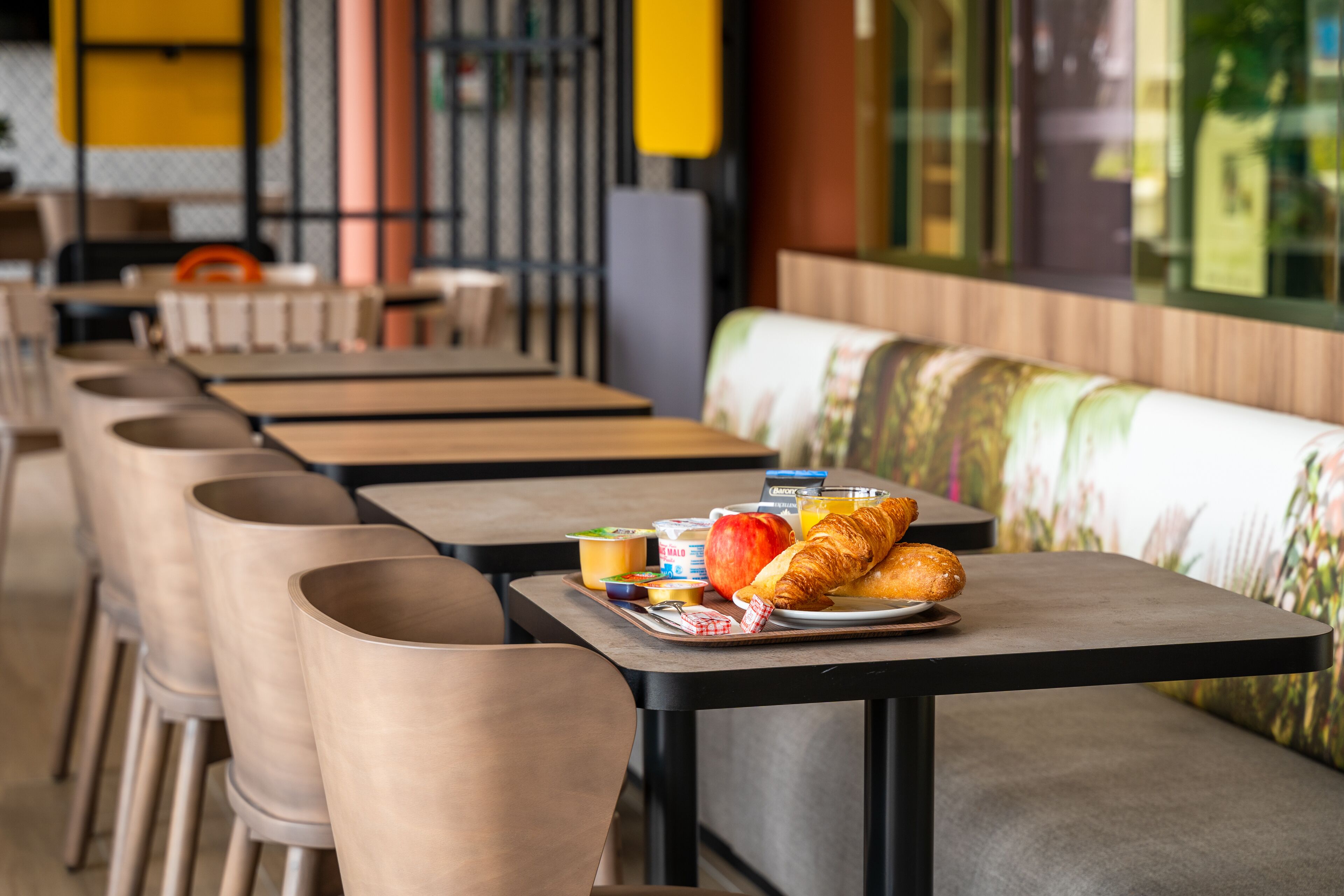 A restaurant with multiple tables and chairs, a tray with croissants, apples, and drinks on one table.