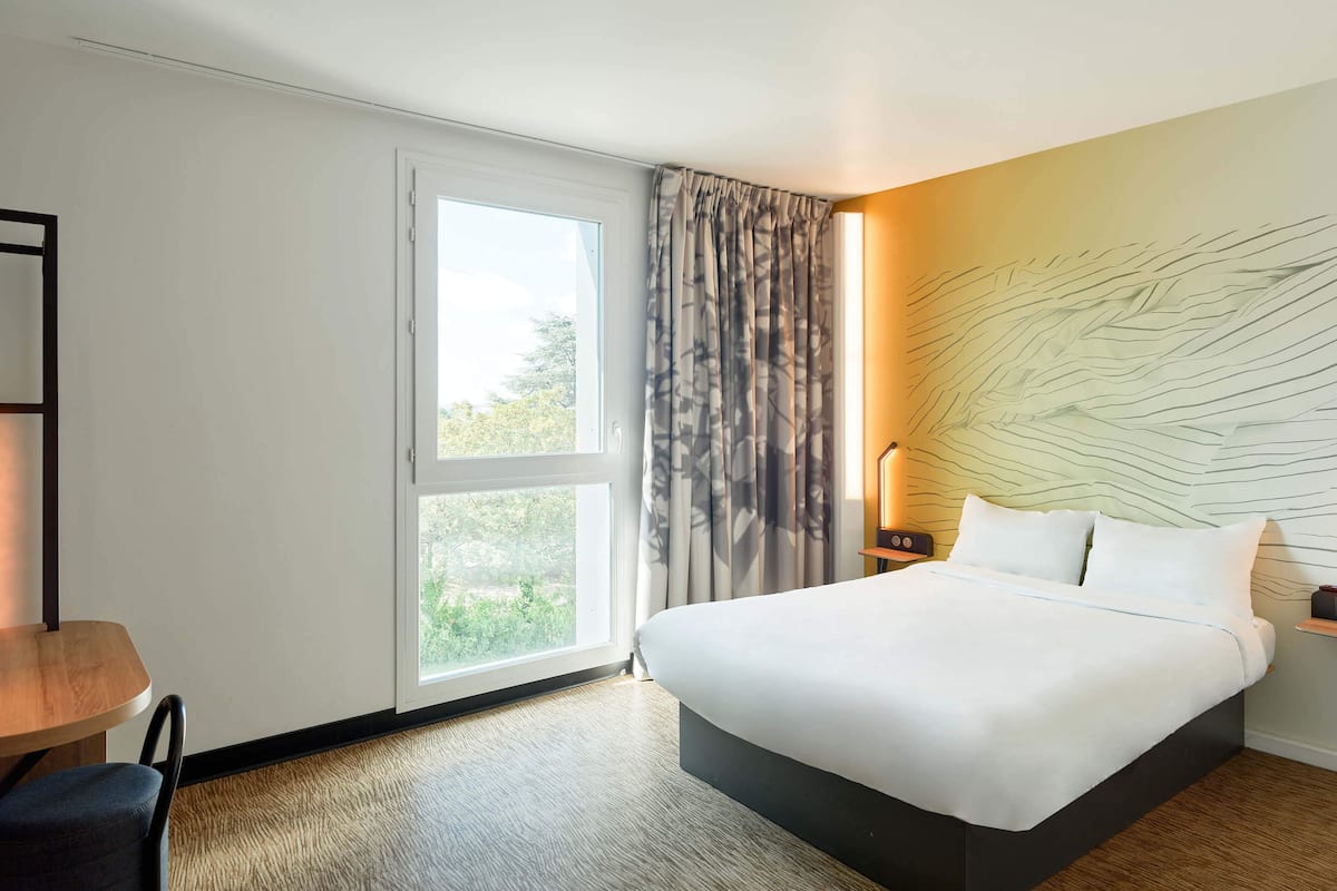 A hotel room with a large bed, a desk, a chair, a window with curtains, and a wall with a textured design.