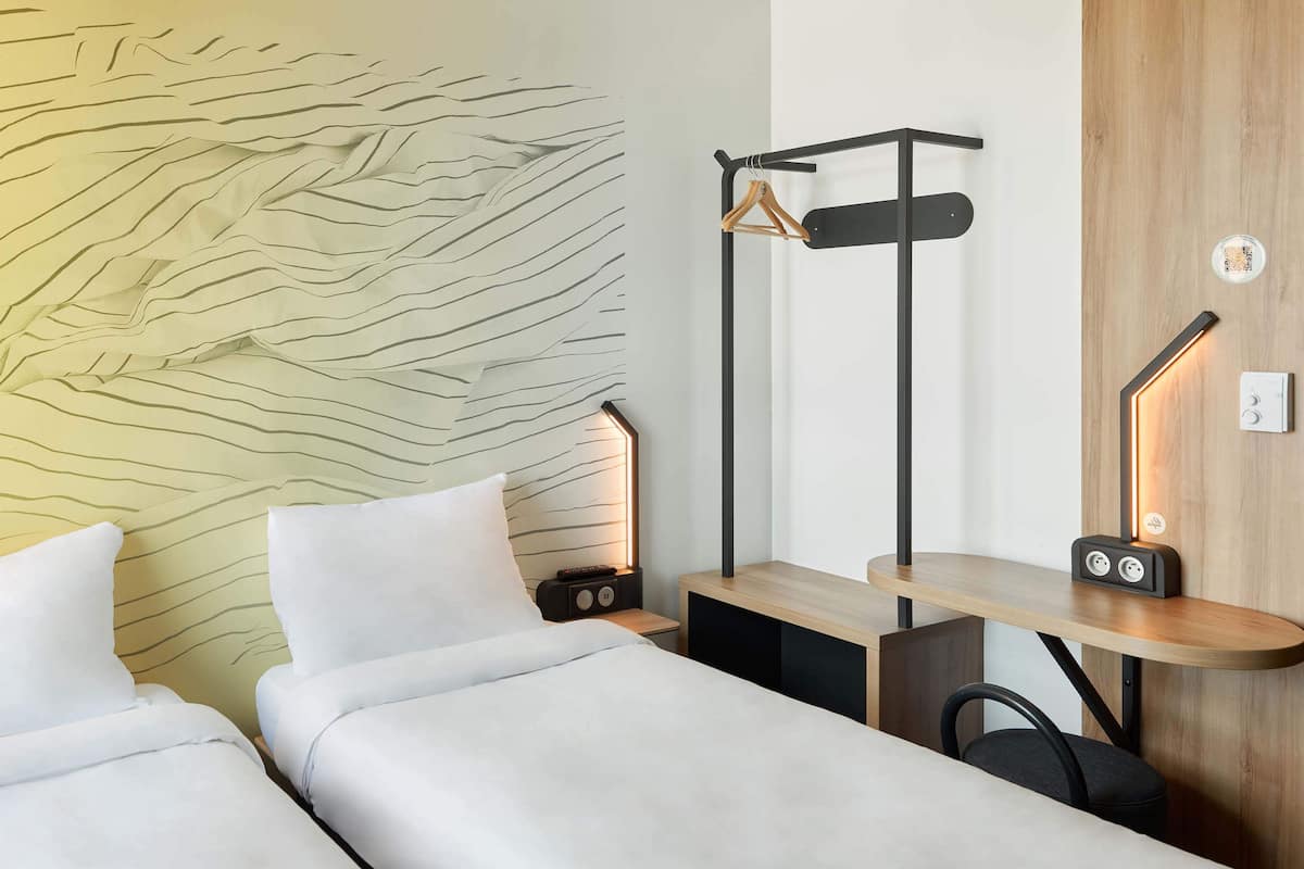 A modern hotel room with two beds, a wooden headboard, a desk with a chair, a coat rack, and bedside lamps.