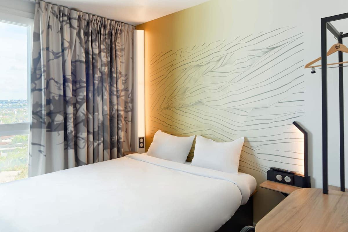 A hotel room with a large bed, a bedside table with a lamp, and a view of the city through the window.