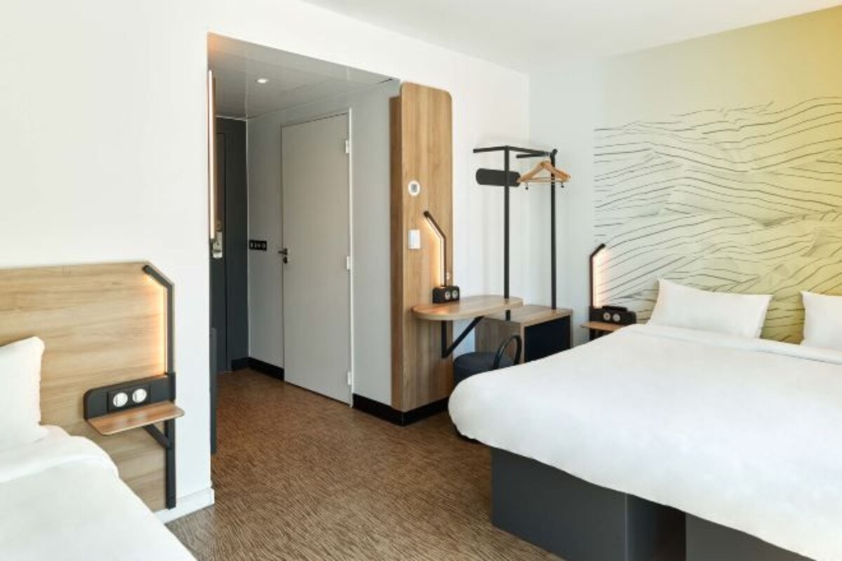 A hotel room with a bed, bedside table, a desk with a chair, a coat rack, and a wall with a textured design.