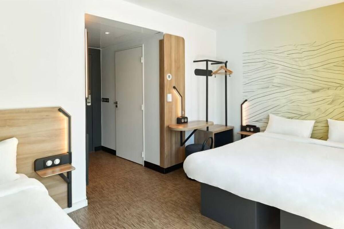 A hotel room with a bed, bedside table, a desk with a chair, a coat rack, and a wall with a textured design.