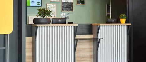 A reception desk with a modern design, featuring a digital screen displaying services and prices, a potted plant, and a clean, striped wall design.