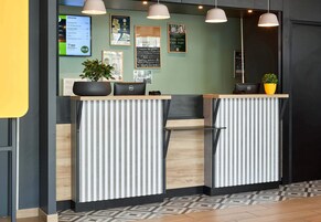 Uma receção com design moderno, apresentando um ecrã digital a mostrar serviços e preços, uma planta em vaso e um padrão de parede listrado e limpo.
