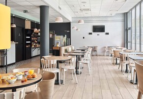 Un café moderno con mesas y sillas de madera, una barra negra con productos para el desayuno, y una pared de ladrillo blanco con un televisor.