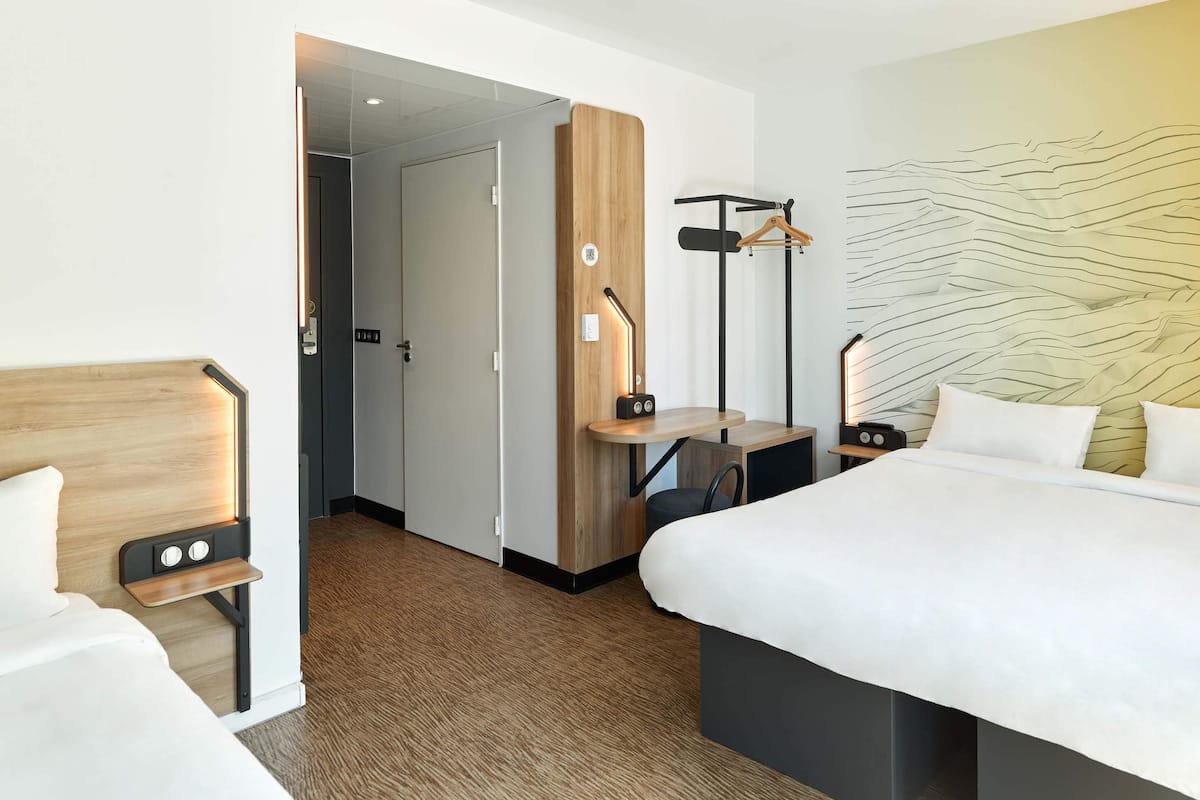 A hotel room with a wooden headboard, a bed with white bedding, a desk with a chair, a wall-mounted coat rack, and a unique textured wall design.