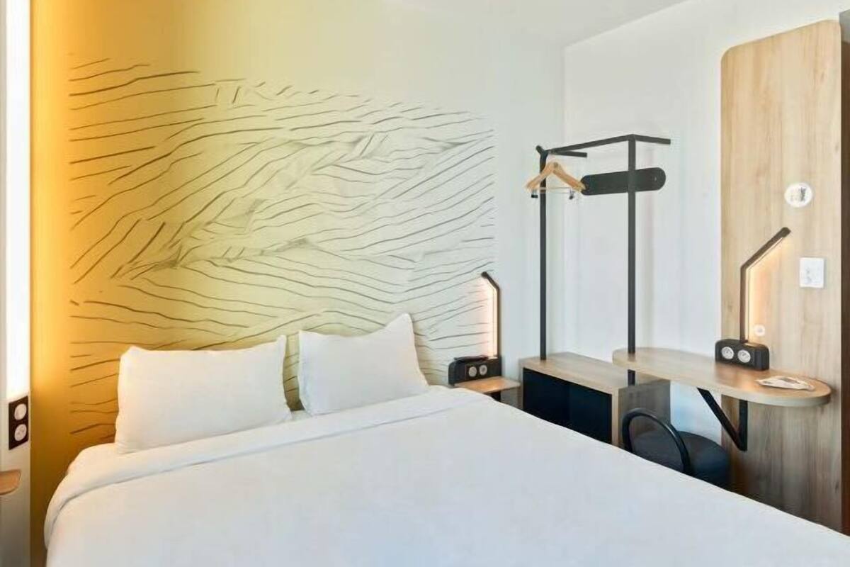 A modern hotel room with a bed, bedside tables, a wooden headboard, and a wall-mounted coat rack.