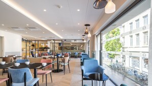 Un café moderno con mesas y sillas, un área de cocina visible y grandes ventanales con vistas a la calle y bicicletas afuera.