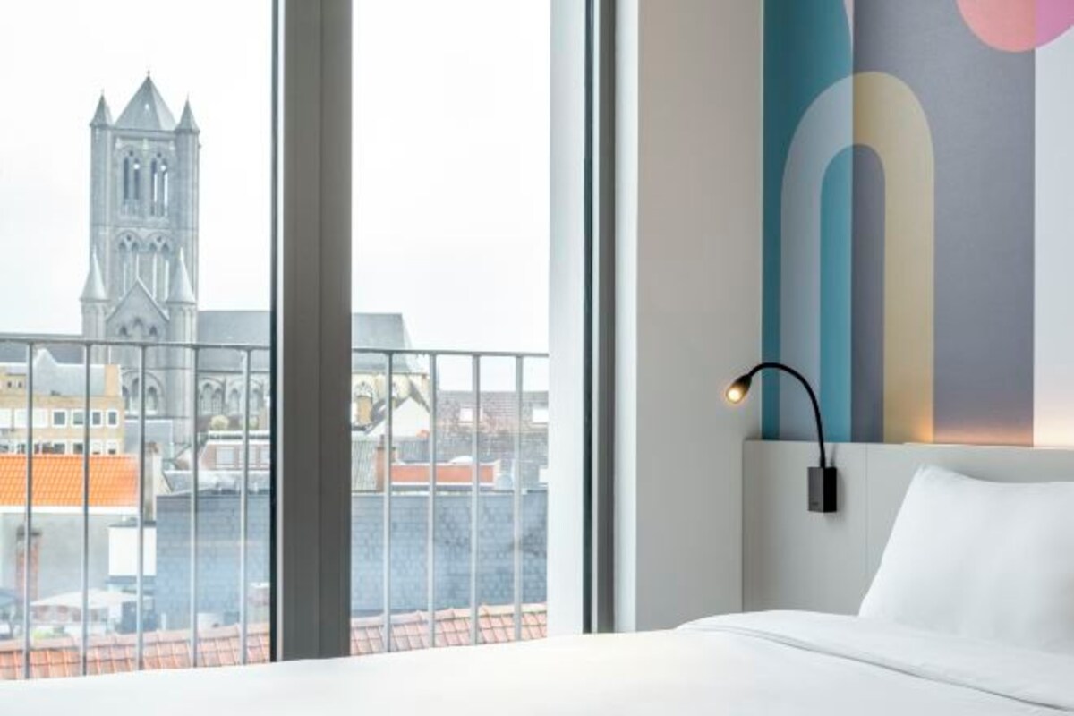 A hotel room with a bed, a nightstand, a wall-mounted lamp, and a large window offering a view of a church tower and cityscape.