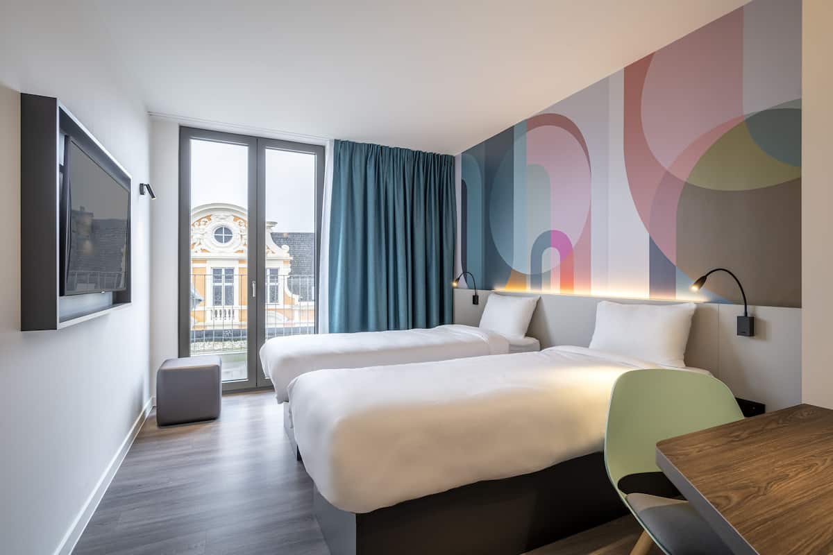 A modern hotel room with two beds, a desk, and a view of a building through a glass door.