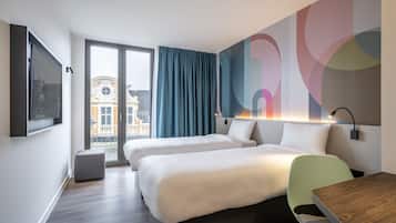 A modern hotel room with two beds, a desk, and a view of a building through a glass door.