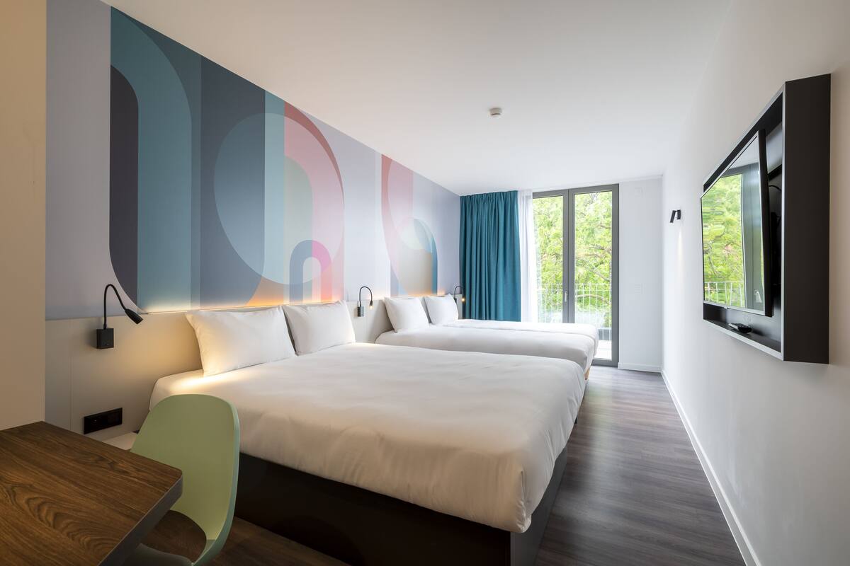 A modern hotel room with two beds, a TV, and a large window with curtains.