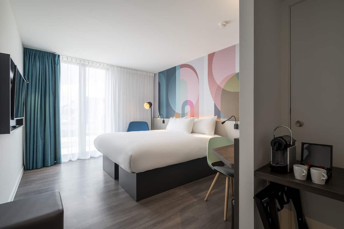 A modern hotel room with a large bed, a desk with a chair, a TV, and a coffee maker.
