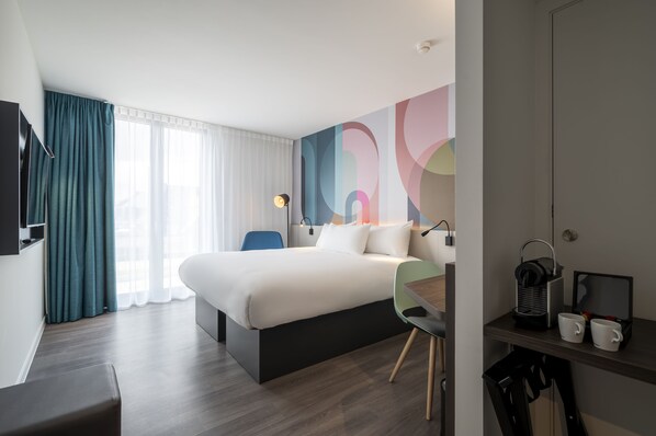 A modern hotel room with a large bed, a desk with a chair, a TV, and a coffee maker.