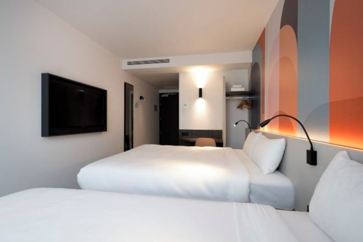 A hotel room with a large bed, a flat-screen TV mounted on the wall, and a bedside table with a lamp.