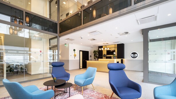 Un moderno vestíbulo de hotel con sillones azules, una recepción y un gran muro de cristal.