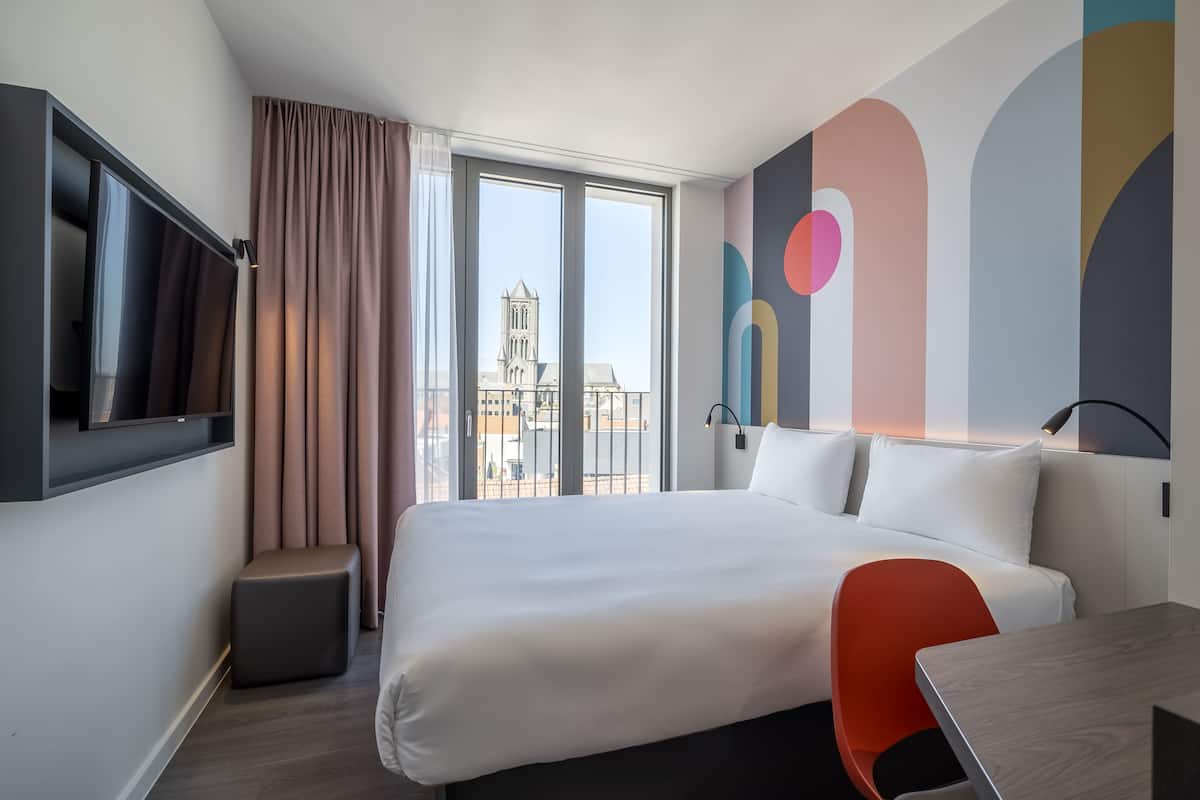 A hotel room with a large bed, a desk, a chair, a TV, and a window with a view of a building.