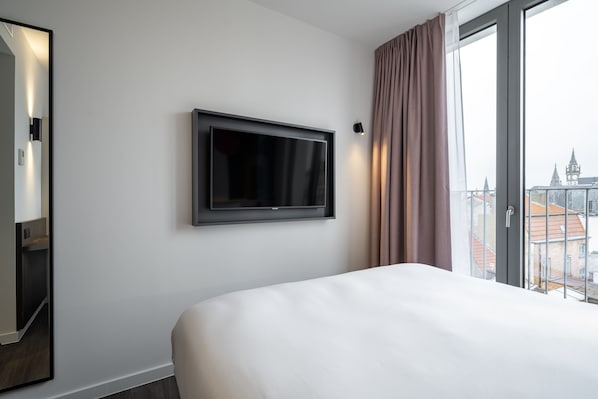 Habitación de hotel con una cama grande, un televisor montado, una ventana con cortinas y vista a la ciudad.
