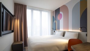Una habitación de hotel moderna con una cama grande, un escritorio y una silla. Hay una ventana con cortinas, un televisor montado en la pared y una pared con arte abstracto.