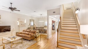A spacious living room with a leather sofa, a coffee table, and a TV. A staircase with a light fixture is visible. - Chic Townhome One Block From Forsyth w/ Large Garage by Lucky Savannah (Savannah)