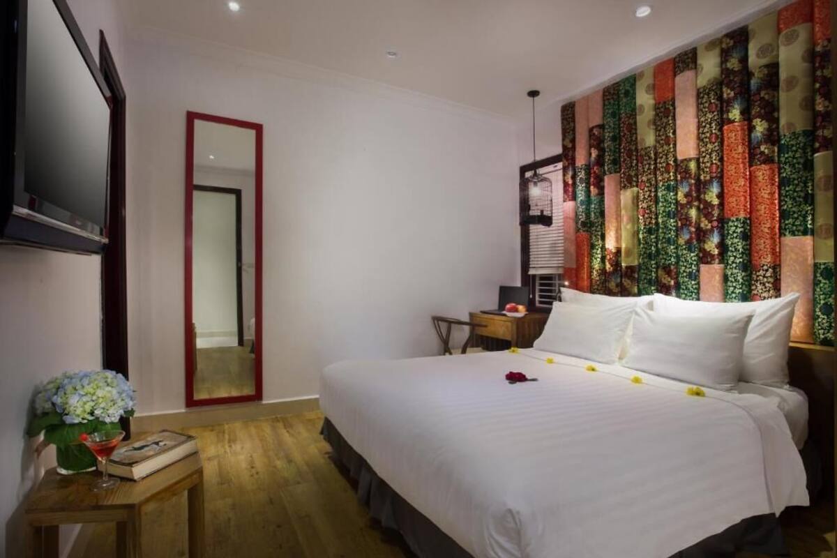 A hotel room with a large bed, a TV mounted on the wall, a wooden nightstand with a vase of flowers, and a colorful wall tapestry.