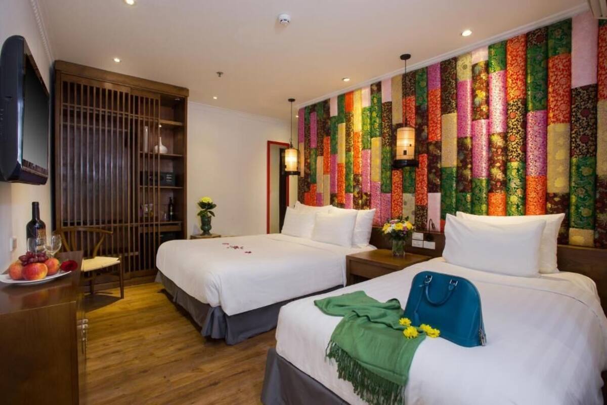 A hotel room with two beds, a wooden floor, a TV, a desk with fruit, and a colorful wall.