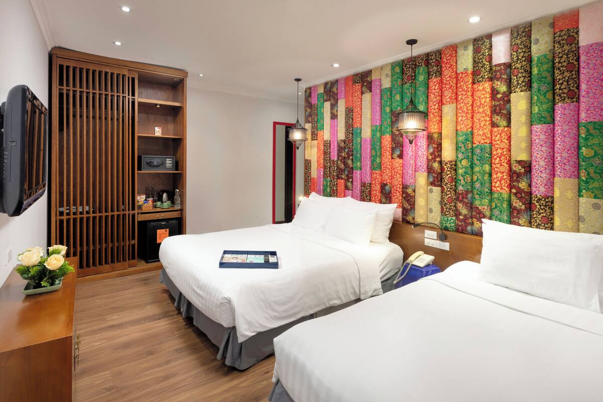 A hotel room with two beds, a TV, a wooden wardrobe, a colorful wall, and a small desk with a vase of flowers.