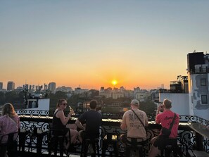 A group of people sitting on a balcony with a cityscape and sunset in the background. - Hanoi Media Hotel And Spa (Hanoi)