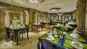 A dining area with ornate columns, chandeliers, and a marble floor. Tables are set with white tableware and green placemats.