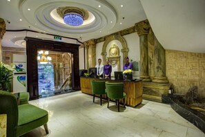 A lobby with a reception desk, green chairs, a chandelier, and a stone wall with columns. - Hanoi Media Hotel And Spa (Hanoi)