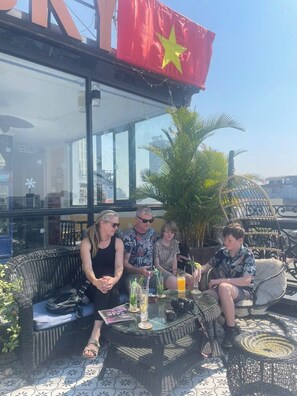 A group of people seated at an outdoor table with drinks, surrounded by wicker furniture and a large potted plant. - Hanoi Media Hotel And Spa (Hanoi)