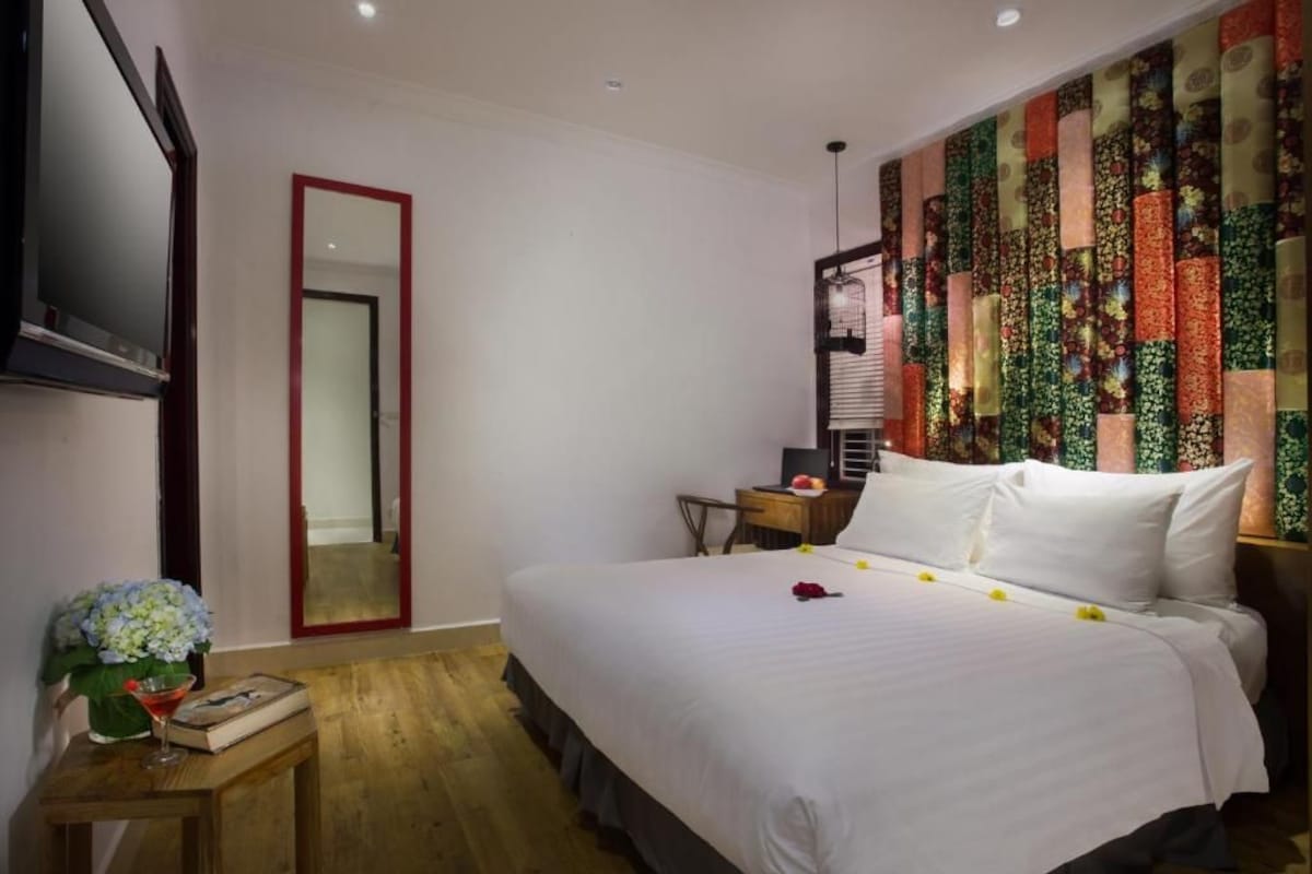 A hotel room with a large bed, a TV mounted on the wall, a wooden nightstand with a vase of flowers, and a colorful wall tapestry.