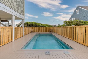 Una piscina rectangular amb una terrassa de fusta, envoltada per una tanca de fusta, i adjacent a un edifici amb façana blanca.