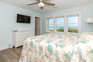 A bedroom with a bed, a ceiling fan, a TV mounted on the wall, and a view of the ocean through the windows.